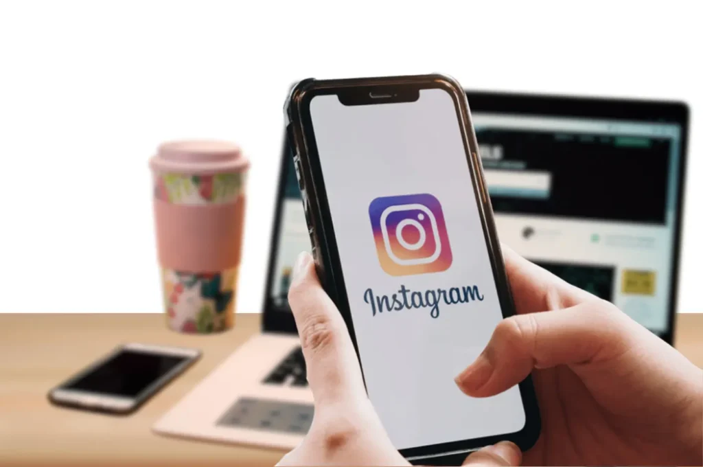 this is the image of a person holding a smartphone using instagra,. behing the phone is a laptop with cofee cup